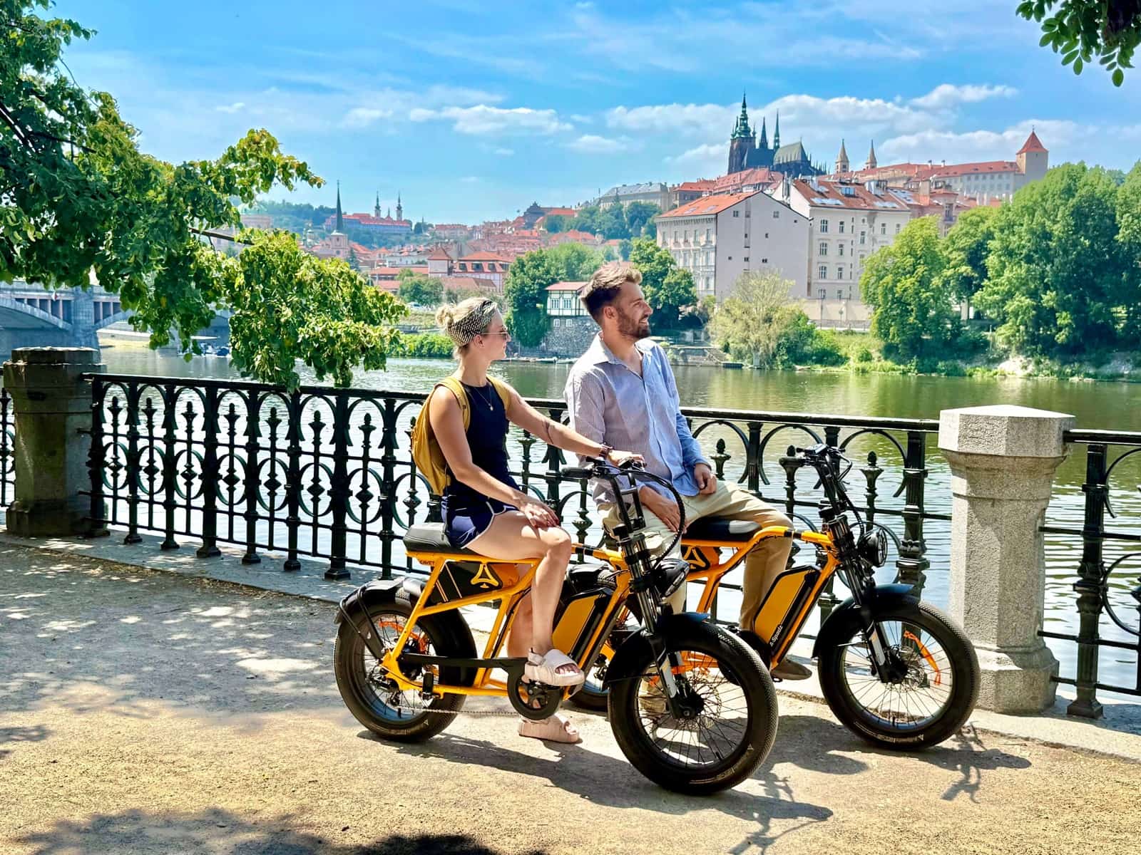 Two riders on orange chopper e-bikes by the river with Prague Castle behind